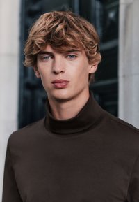 Brun polotröja i ett mjukt material med figurnära passform och hög krage. Närbild på en ung man mot en suddig bakgrund.