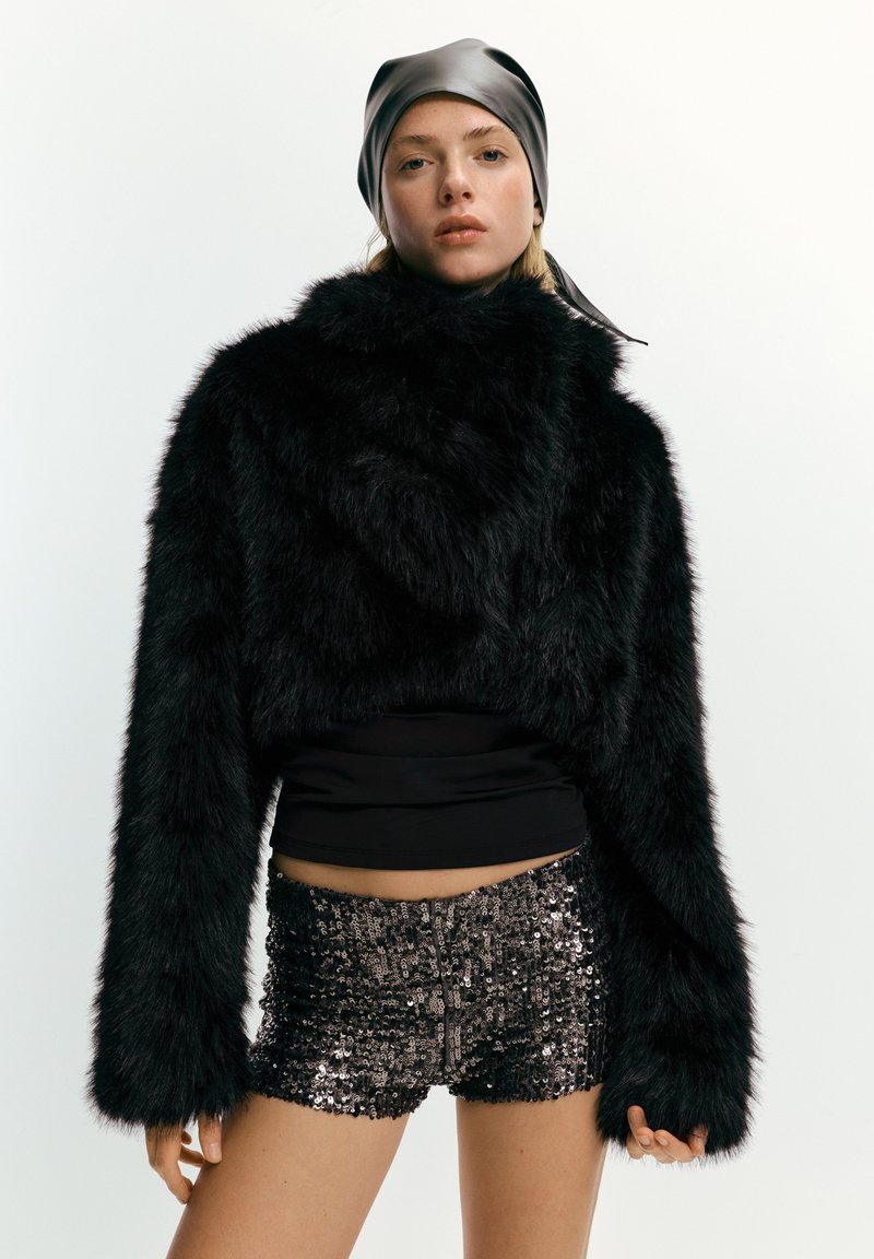 Musta tekoturkistakki, jossa on leveät hihat ja lyhyt pituus, yhdistetty paljettishortseihin. Mallilla on hopeinen satiininen päähuivi. Minimalistinen tausta.