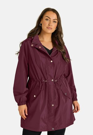 Femme aux longs cheveux portant un imperméable bordeaux avec des boutons sur le devant, une taille à cordon et des poches à rabat, debout devant un fond uni.