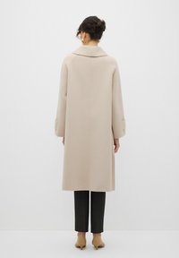 Cappotto color beige in misto lana con ampio colletto e spalle scese, lungo fino al ginocchio, abbinato a pantaloni neri a righe e scarpe beige.