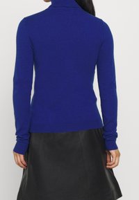 Blå polotröja i mjukt tyg, med långa ärmar och ribbade mudd och nederkant. Bärs tillsammans med en svart kjol.