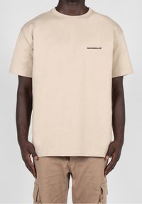 Beige bavlněné tričko s krátkým rukávem a kulatým výstřihem, s malým černým logem na hrudi, nošené s světle hnědými Cargo kalhotami.