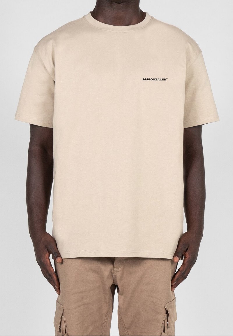 Beige bavlněné tričko s krátkým rukávem a kulatým výstřihem, s malým černým logem na hrudi, nošené s světle hnědými Cargo kalhotami.