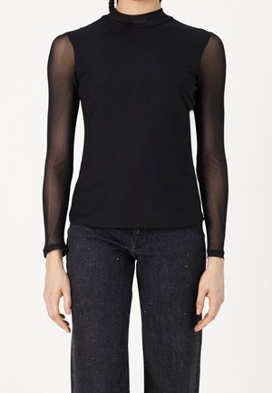 Personne portant un haut noir moulant avec des manches longues transparentes et un pantalon noir aux détails subtilement mouchetés, debout les bras détendus.