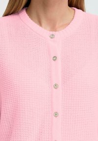Rosa stickad kofta med ribbad halsringning, prydd med två rader dekorativa silverfärgade knappar fram.