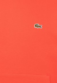 Tissu orange vif avec un petit logo de crocodile vert brodé près du centre supérieur et une couture sur le bas.