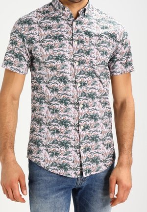 Hombre que lleva una camisa de manga corta con botones y estampado de palmeras verdes y flamencos, combinada con vaqueros azules.