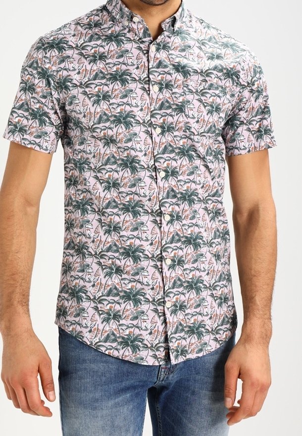 Homme portant une chemise à manches courtes boutonnée avec un imprimé de palmiers verts et de flamants roses, associée à un jean bleu.
