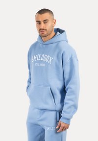 Sudadera azul celeste con capucha, que presenta un bolsillo delantero y el texto blanco "SMILODOX ATHL. WEAR". La tela parece suave y texturizada.