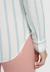 White long-sleeve shirt with light blue vertical stripes, featuring buttoned cuffs and a curved hem, paired with pink pants.