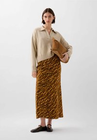 Pull beige court avec un décolleté en V, associé à une jupe jaune moutarde ornée d'un imprimé animal sombre. Le mannequin tient une pochette marron.