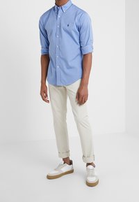 Muž v světlemodré košili s dlouhým rukávem a ohrnutými manžetami, béžových kalhotách ohrnutých nad kotníky a bílých teniskách s gumovými podrážkami.