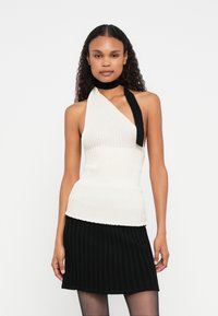 Top halter assimétrico branco canelado com um detalhe de laço preto ao redor do pescoço, combinado com uma saia curta preta plissada.