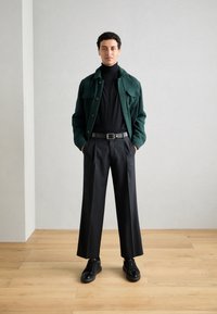 Vihreä corduroy-takki napitettuna edessä, musta poolopaita, mustat housut ja teksturoidut mustat kengät. Yksinkertainen muotoilu modernilla istuvuudella.