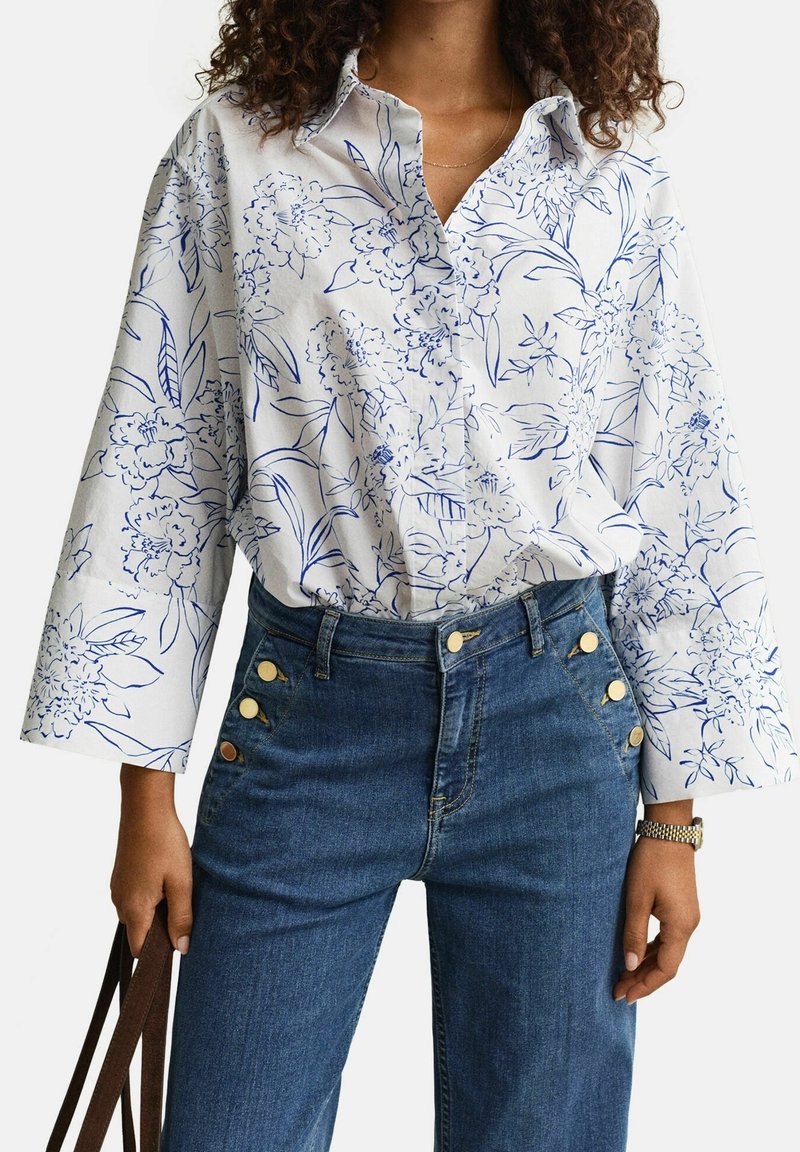 Femme portant une chemise blanche boutonnée avec un imprimé floral bleu et un jean bleu taille haute avec des boutons dorés, tenant des anses de sac marron.