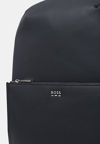 Sac à dos noir texturé avec une poche zippée à l'avant et des accessoires en métal argenté. Présente le nom de la marque "BOSS" imprimé de manière proéminente.