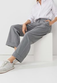 Femme assise sur un bloc blanc portant une chemise blanche, un pantalon large à fines rayures grises et des baskets blanches.