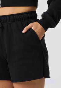 Person trägt schwarze Shorts mit elastischem Bund und Hand in der Tasche, kombiniert mit einem schwarzen, kurz geschnittenen Sweatshirt, ein Ärmel ist sichtbar.