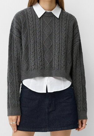 Frau trägt einen grauen, kurz geschnittenen Pullover mit Zopfmuster über einem weißen Hemd mit Kragen und einem dunklen Jeans-Minirock, steht mit entspannten Händen.