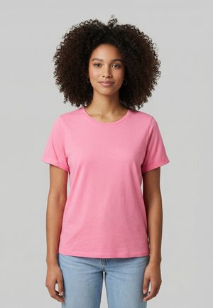 Giovane donna con capelli ricci che indossa una maglietta rosa a maniche corte e jeans azzurri, in piedi davanti a uno sfondo grigio uniforme.