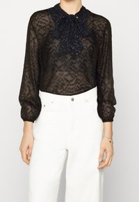 Blusa nera trasparente con motivo testurizzato, maniche lunghe a sbuffo e dettaglio del colletto con fiocco, abbinata a jeans bianchi a vita alta.