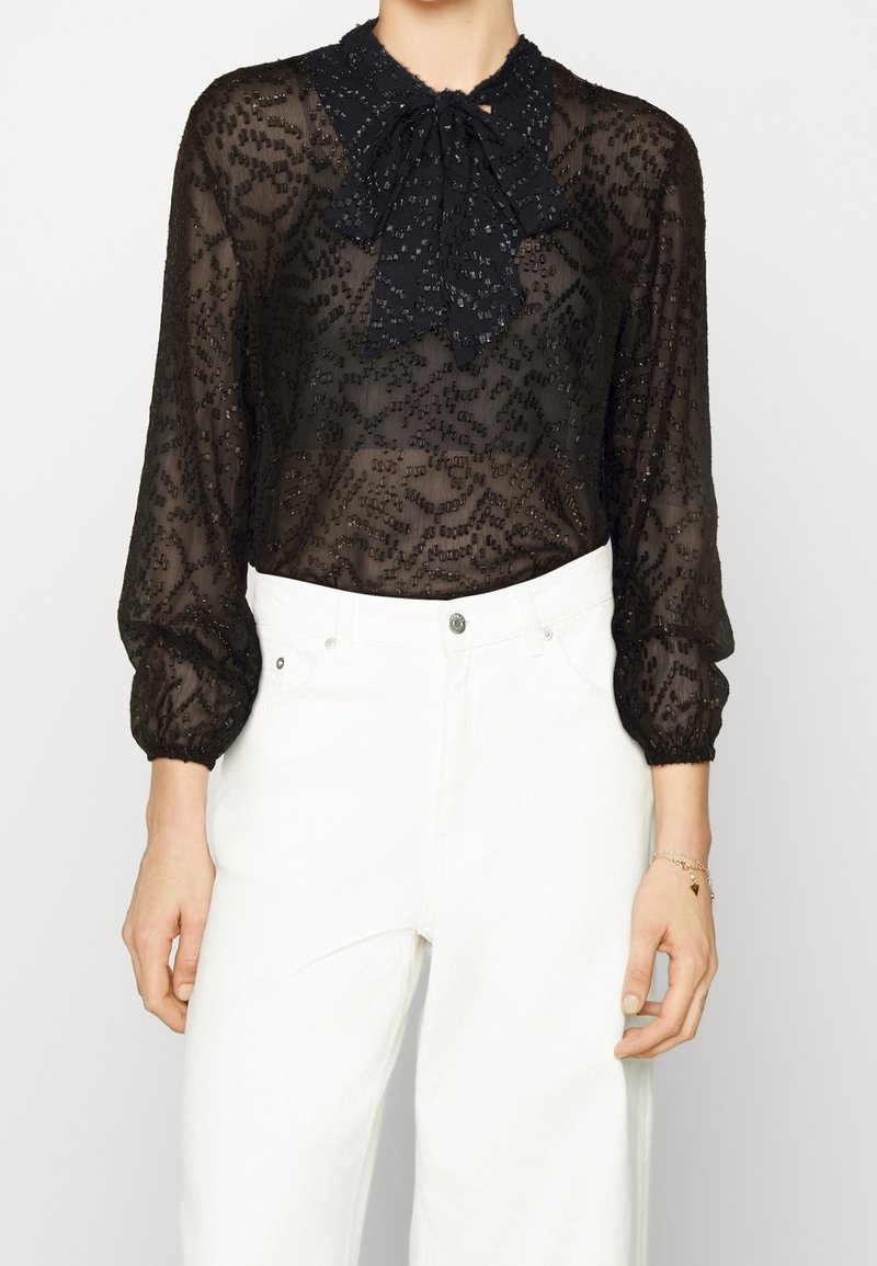 Blusa nera trasparente con motivo testurizzato, maniche lunghe a sbuffo e dettaglio del colletto con fiocco, abbinata a jeans bianchi a vita alta.