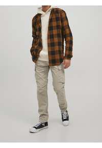 Brun och svart rutmönstrad flanellskjorta över en vit hoodie, tillsammans med beigefärgade cargobyxor och svarta sneakers med vita sulor.