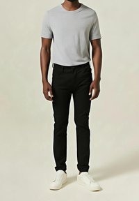 Homme portant un t-shirt gris clair à manches courtes, un pantalon noir slim et des baskets blanches, debout devant un fond uni.