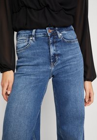 Vaqueros de mezclilla azul de cintura alta con cinco bolsillos, piernas rectas y un ligero desgaste, combinados con una blusa negra semitransparente.