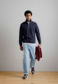 Pullover blu navy con zip, maglietta bianca, jeans azzurri chiari, in mano una giacca bordeaux, indossando sneakers grigie e rosse, che cammina su un pavimento di legno.