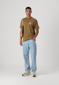 Camiseta de algodón marrón con un pequeño logo en forma de triángulo, jeans de mezclilla azul claro, zapatillas beige y un bolso de hombro marrón. Estilo simple y de corte relajado.