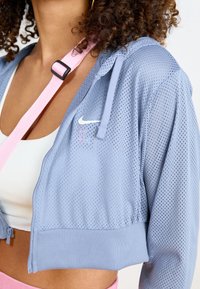 Ljusblå mesh zip-up jacka med en åtsittande midja, prydd med en vit Nike-logotyp och ett rosa band över axeln.