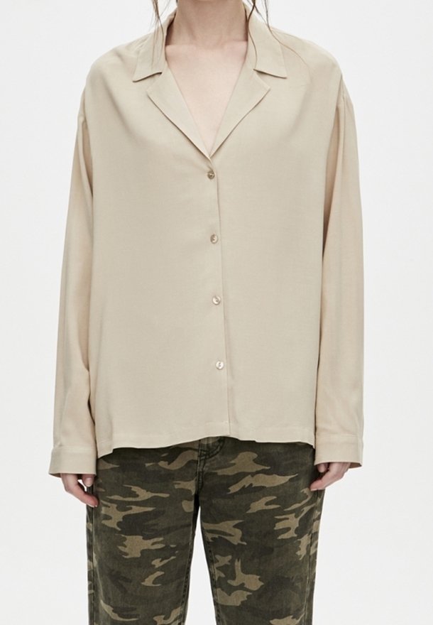 Personne portant une chemise beige ample à boutons et un pantalon de camouflage vert, debout devant un fond blanc uni.