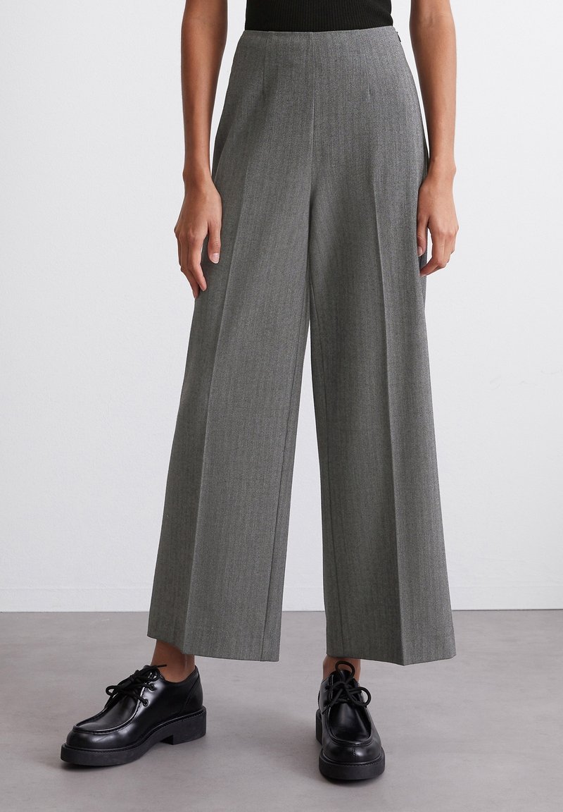Femme portant un pantalon gris taille haute à jambes larges, associé à des chaussures en cuir noires à lacets, debout sur un sol uni contre un mur blanc.