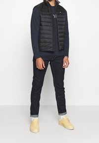 Czarna pikowana kamizelka bez rękawów na granatowej koszuli z długim rękawem, w połączeniu z ciemnoniebieskimi dżinsami i żółtymi trampkami. Białe skarpetki widoczne przy kostkach.