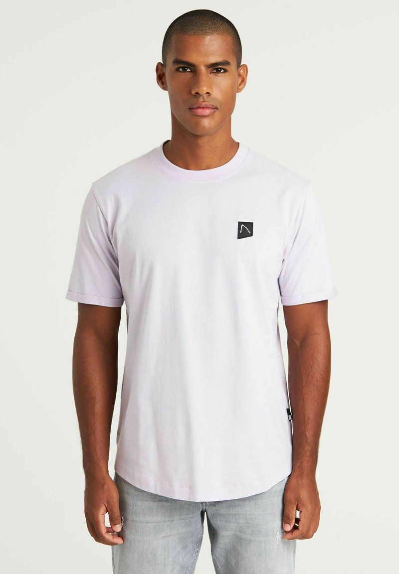 Liliowa koszulka z krótkim rękawem wykonana z bawełny, z małą czarną naszywką z logo na lewej piersi i prostym dołem. Noszona z jasnoszarymi dżinsami.