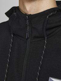 Schwarze Zip-Hoodie mit hohem Kragen, verstellbaren Kordeln und strukturiertem Stoff. Besitzt dezente reflektierende Akzente und ein Logo-Patch.