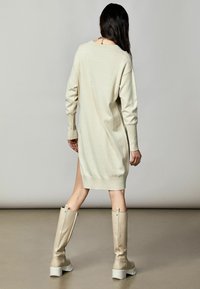 Vestido de punto crema con mangas largas, aberturas laterales y puños acanalados, combinado con botas beige hasta la rodilla con suela blanca gruesa.