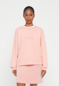 Sweatshirt oversized rosa com gola e barra caneladas, apresentando um logótipo da marca bordado. Combinado com uma saia de sarja correspondente.
