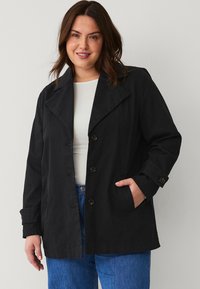 Zwarte, knopen-up jas met grote kraag, zijzakken en opgerolde mouwen, gemaakt van een gestructureerde stof, gecombineerd met een witte top en blauwe jeans.