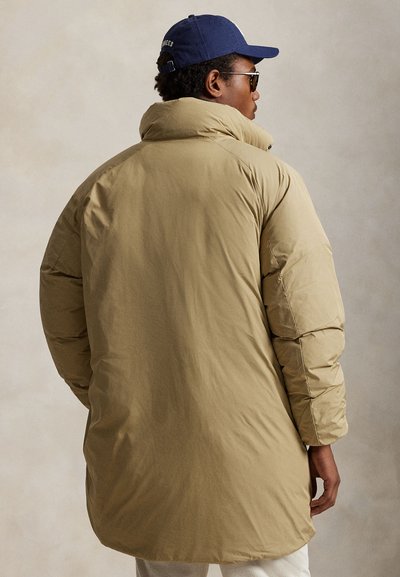 Bēša pūkaina jaka ar augstu apkakli, plašu piegriezumu un bieza polsterējuma slāni. Raksturo gluda tekstūra un viegli noapaļota muguras mala.