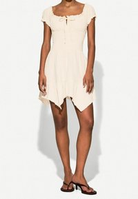 Kurzes beige Kleid mit gesmoktem Oberteil, Rüschensäumen, die die Schultern freilassen, und einer asymmetrischen Saumtextur. Getragen mit schwarzen, geschnallten Sandalen.