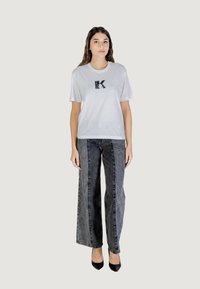 T-shirt bianca con logo nero, abbinata a jeans grigi a gamba larga. La modella indossa tacchi alti neri. Design semplice con vestibilità casual.