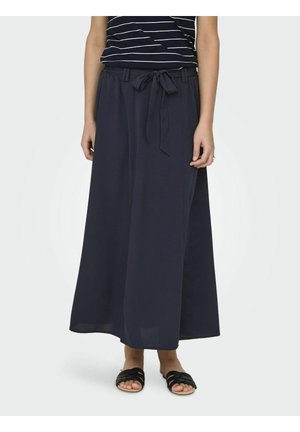 Woman wearing a navy blue wide-leg skirt with a tied waist belt, striped top, and black open-toe flat sandals, standing on a light background.