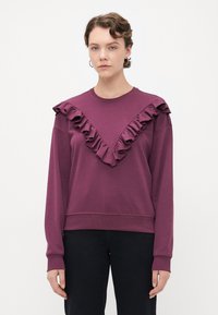 Giovane con un maglione viola decorato con dettagli a volute a forma di V sul davanti e pantaloni neri, su uno sfondo bianco uniforme.