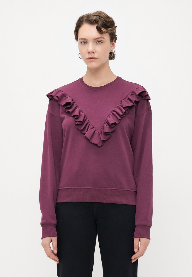 Giovane con un maglione viola decorato con dettagli a volute a forma di V sul davanti e pantaloni neri, su uno sfondo bianco uniforme.