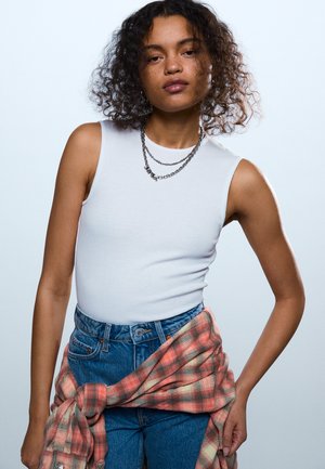 Mladá žena s kučeravými vlasmi má na sebe biely tielkový top, vrstvené strieborné reťaze, modré džínsy a okolo pása zviazanú červenú károvanú košeľu.