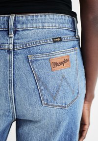 Plave traperice imaju smeđi kožni detalj s "Wrangler" logotipom. Dizajn s pet džepova sa vidljivim šavovima i prepoznatljivim W uzorkom na stražnjem dijelu.
