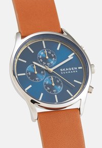 Orologio Skagen con quadrante blu, cassa argentata, tre sotto-quadranti e accenti dorati. Presenta un bracciale in pelle marrone chiaro con una texture liscia.