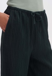 Donkergroene, gestructureerde, geplooide broek met een elastische tailleband en trekkoord. Zakken aan de zijkanten zijn zichtbaar langs de naden.